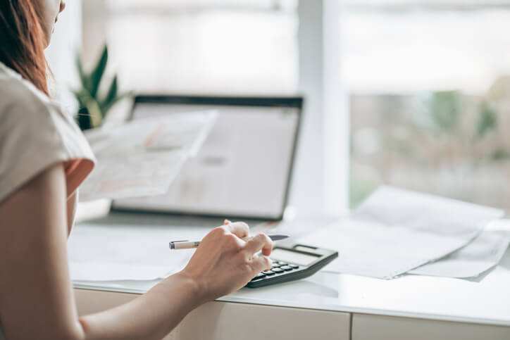 person using calculator on a desk with their laptop in the background