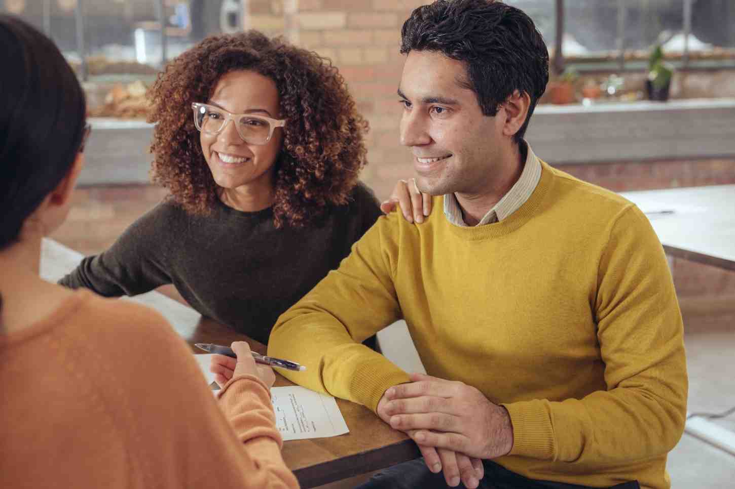 two people sitting at a table across from another person who is helping them with paperwork