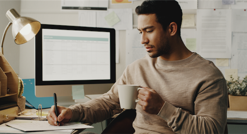 person writing down a budget with computer in background and a coffee in hand